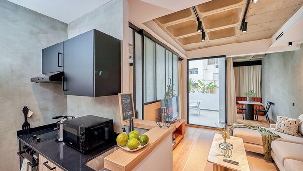 Una cocina moderna con microondas integrado, un lavamanos y una encimera con frutas. Hay una zona de comedor con un sofá y una mesa de centro.