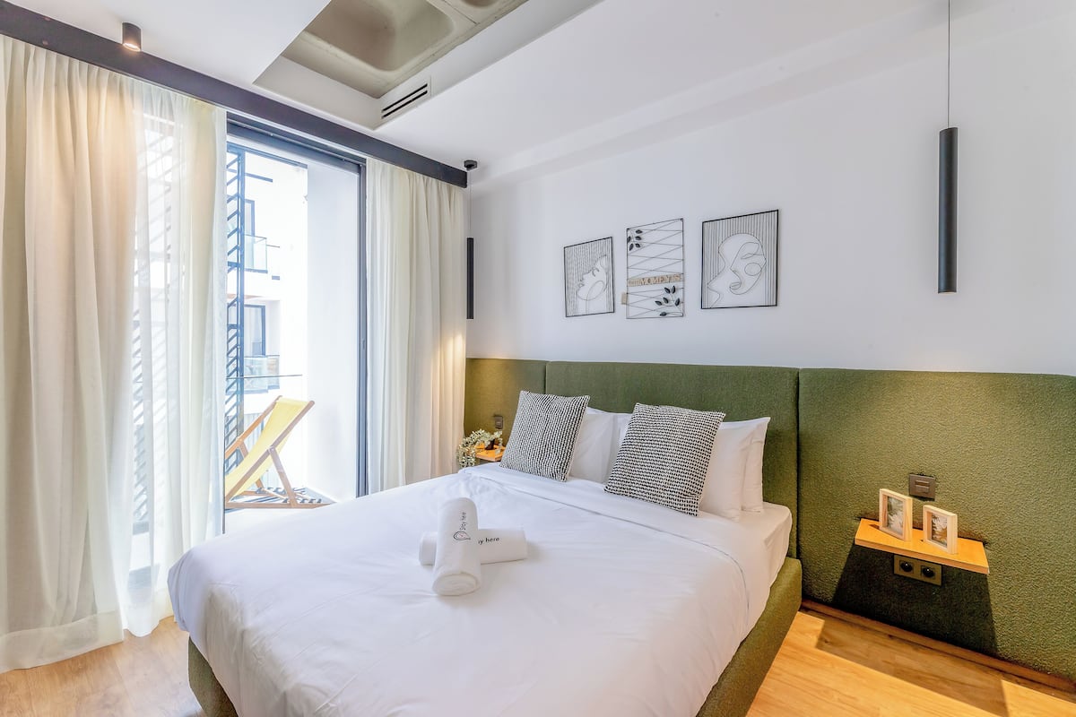 A neatly made bed with a white comforter, pillows, and a rolled towel. There are framed artworks on the wall, a nightstand with a lamp, and a view of the outside through a window with curtains.