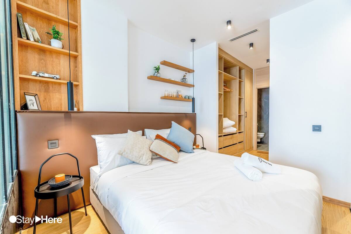 A modern hotel room with a neatly made bed, a bedside table with a lamp, and built-in wooden shelving with books and decorative items.