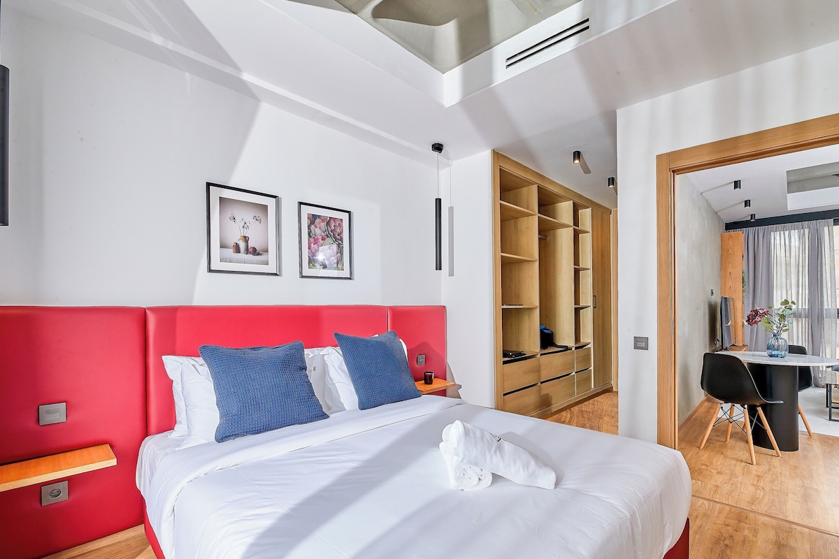 A modern hotel room with a red headboard, white bedding, and a neatly made bed with a towel. There is a wooden wardrobe, a desk with a chair, and a large window with curtains.