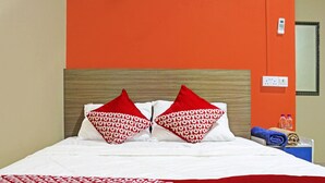 A hotel room with a bed, two red pillows with a pattern, a white blanket, a red and white patterned bedspread, a wooden headboard, a wall socket, and a mirror.