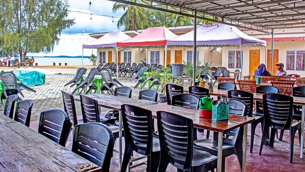 A beachside restaurant with outdoor seating, covered by a metal roof, and overlooking the sea.