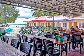 A beachside restaurant with outdoor seating, covered by a metal roof, and overlooking the sea.