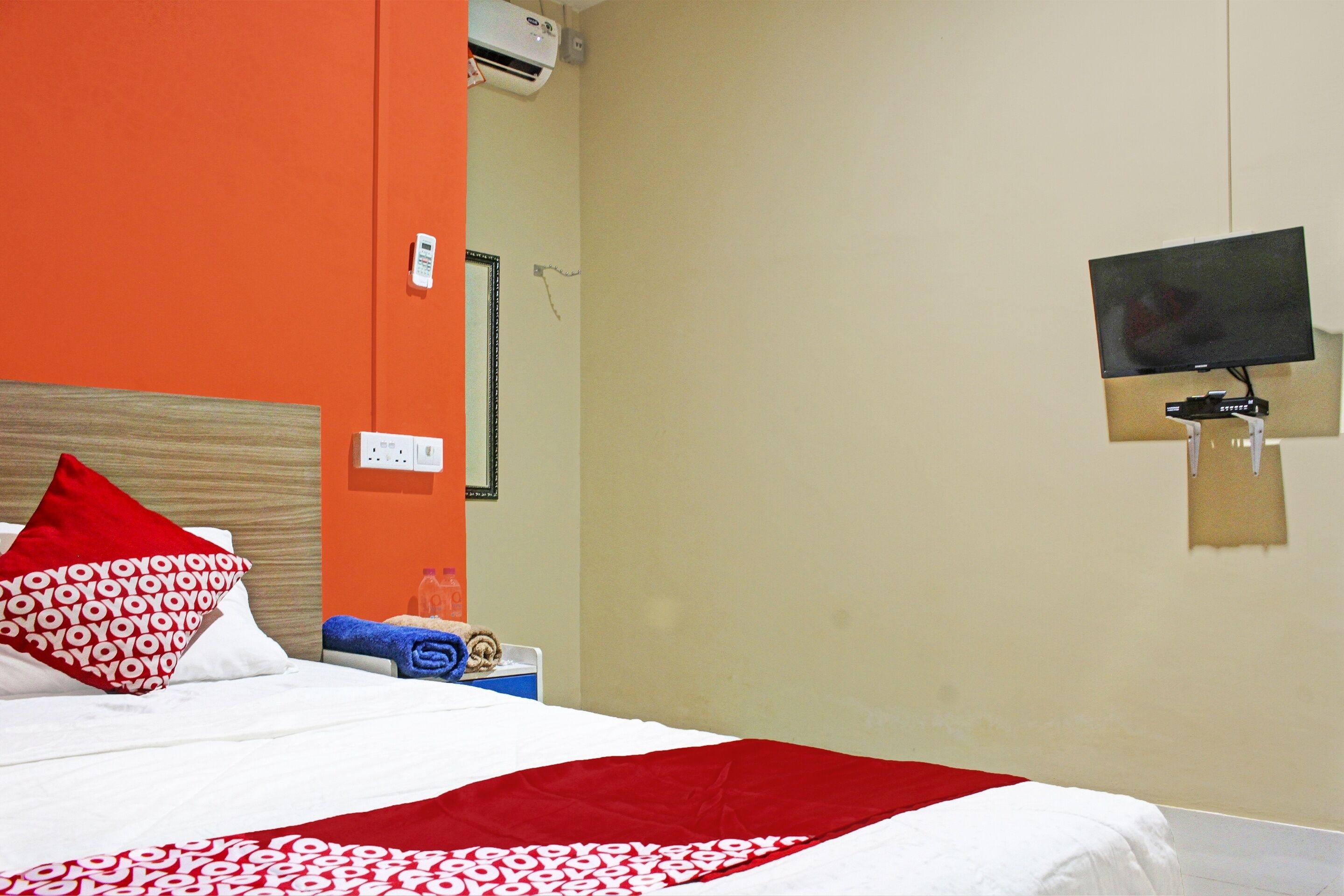 A hotel room with a bed, a red and white patterned pillow, a red blanket, a television mounted on the wall, and a small table with a bottle and a tissue box.
