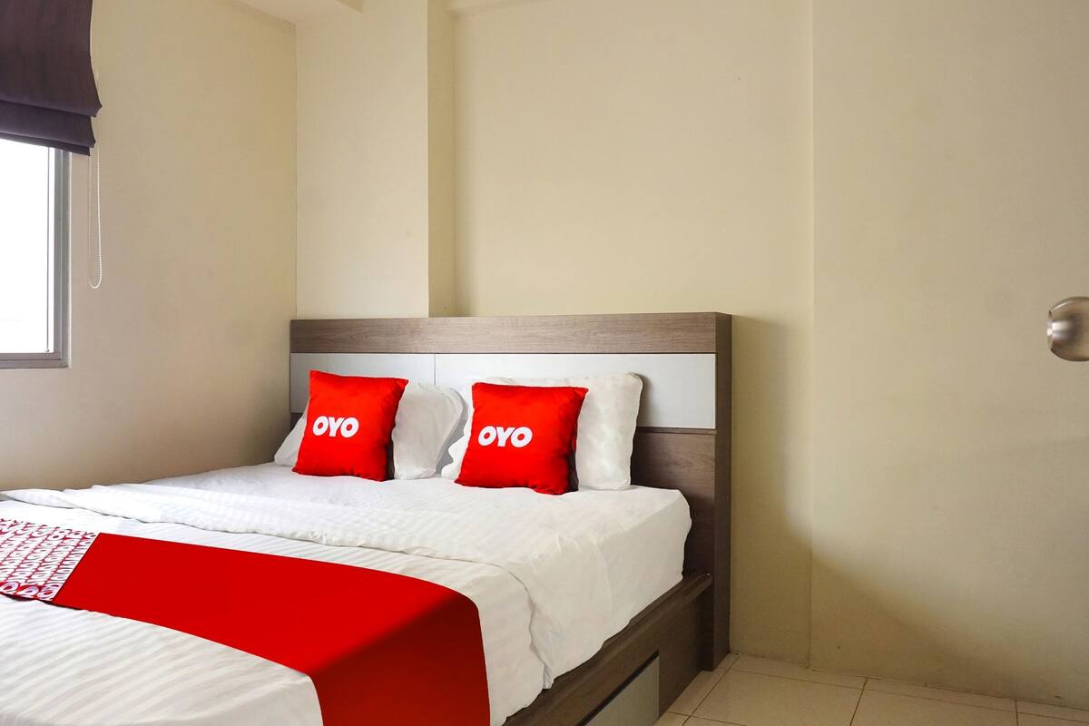 A neatly made bed with white bedding and two red pillows with "OYO" text, a wooden headboard, and a window with a dark curtain.