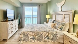 A bedroom with a bed, a television on a dresser, a ceiling fan, a nightstand with a lamp, and a view of the ocean through a sliding door.