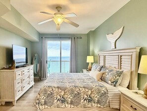 A bedroom with a bed, a television on a dresser, a ceiling fan, a nightstand with a lamp, and a view of the ocean through a sliding door.