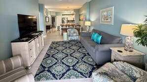 A living room with a sofa, a TV, a patterned rug, and a dining area in the background.