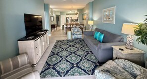 A living room with a sofa, a TV, a patterned rug, and a dining area in the background.