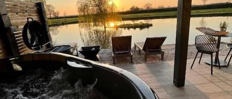 A hot tub with a view of a sunset over a body of water, outdoor seating, and a covered patio area.