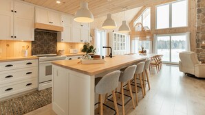Una cocina moderna con armarios blancos, una isla grande con taburetes de bar, iluminación colgante y vistas a un paisaje nevado a través de amplios ventanales.
