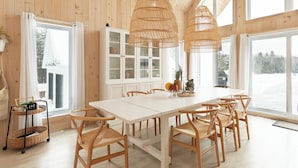 Un comedor amplio con una mesa blanca y sillas de madera, ventanas grandes que ofrecen vistas al exterior, y un mueble blanco con puertas de vidrio.