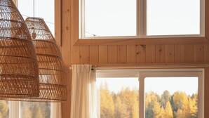 Un comedor con mesa y sillas de madera, ventanas amplias con vista a un bosque, y una lámpara de techo trenzada.