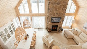 Una habitación acogedora y bien iluminada, con chimenea de piedra, paredes de madera y ventanas amplias que ofrecen vistas a un paisaje nevado.
