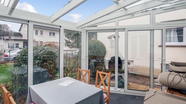 Una veranda con tavolo e sedie, ampie porte in vetro e vista sul giardino e sulle case vicine.