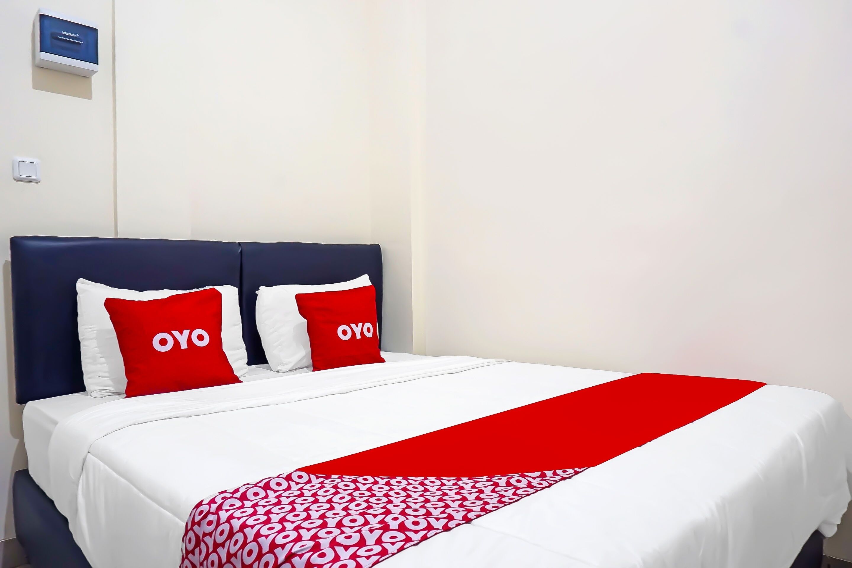 Una habitación de hotel con una cama, dos almohadas con la palabra 'OYO', una colcha blanca y una manta estampada en rojo y blanco.