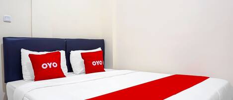 Una habitación de hotel con una cama, dos almohadas con la palabra 'OYO', una colcha blanca y una manta estampada en rojo y blanco.