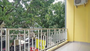 Un balcon avec un mur jaune, une balustrade blanche et une vue sur une rue bordée d’arbres et où sont garées des voitures.