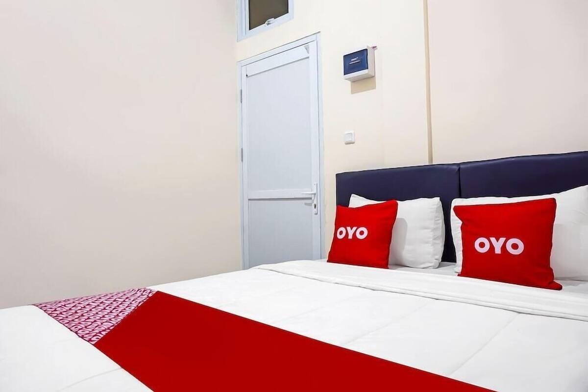 Een hotelkamer met een bed, twee rode kussens met de tekst 'OYO', een witte deur en een klein blauw bord aan de muur.