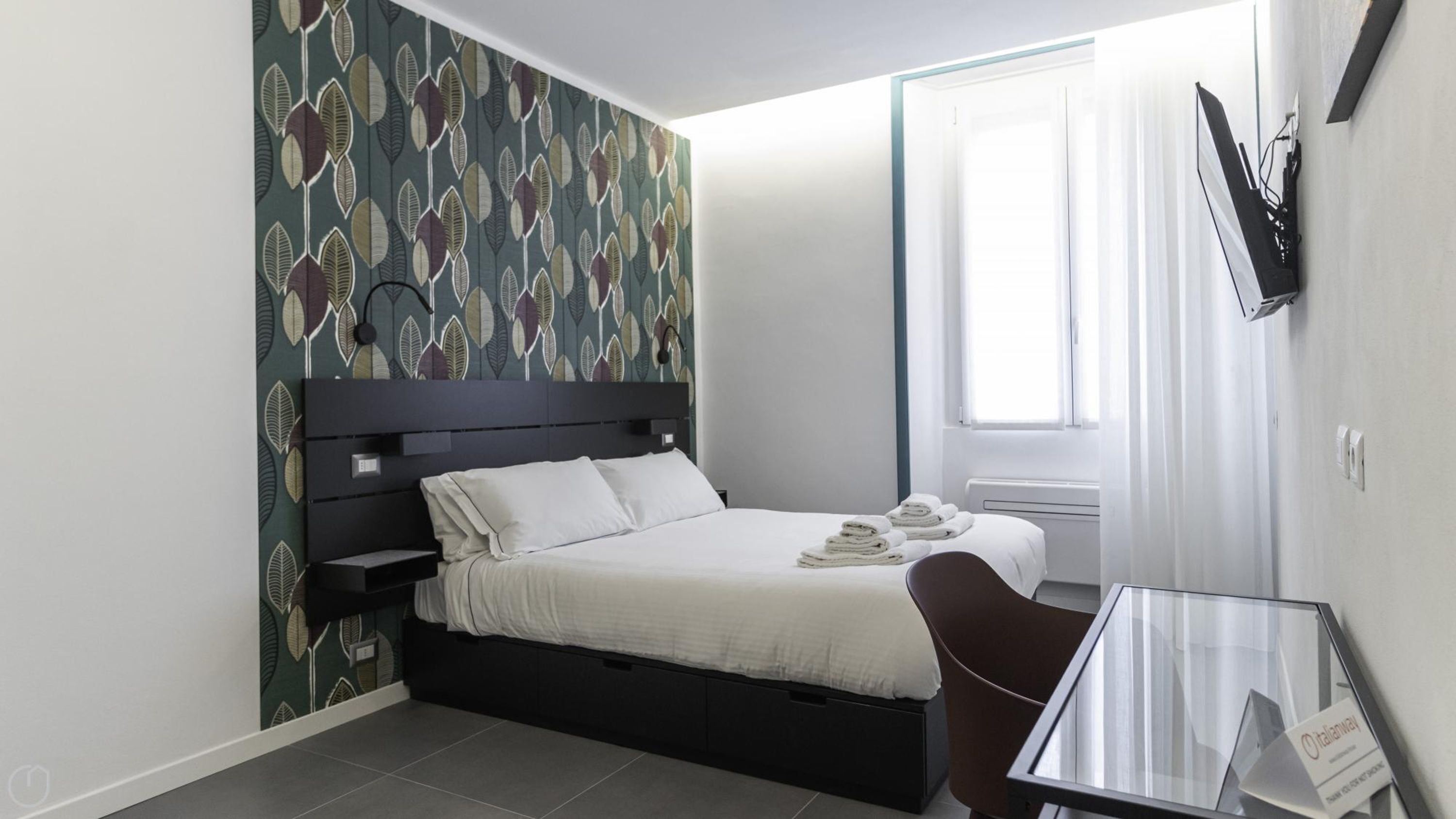 Ein modernes Hotelzimmer mit Bett, Schreibtisch, Stuhl, Wandfernseher und einem Fenster mit Vorhängen.