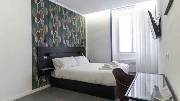 A modern hotel room with a bed, a desk, a chair, a TV mounted on the wall, and a window with curtains.