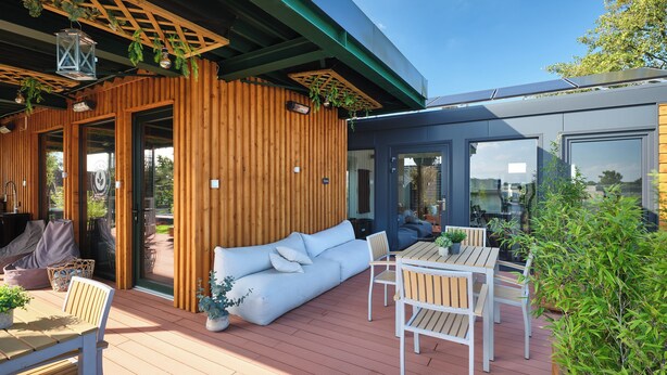 A wooden deck with a sofa, chairs, and a table, a glass door leading to a room, and solar panels on the roof. - PHIL & MAX Hotel & Apartments  Airport (Langenhagen)