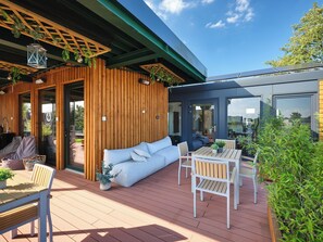 A wooden deck with a sofa, chairs, and a table, a glass door leading to a room, and solar panels on the roof. - PHIL & MAX Hotel & Apartments  Airport (Langenhagen)
