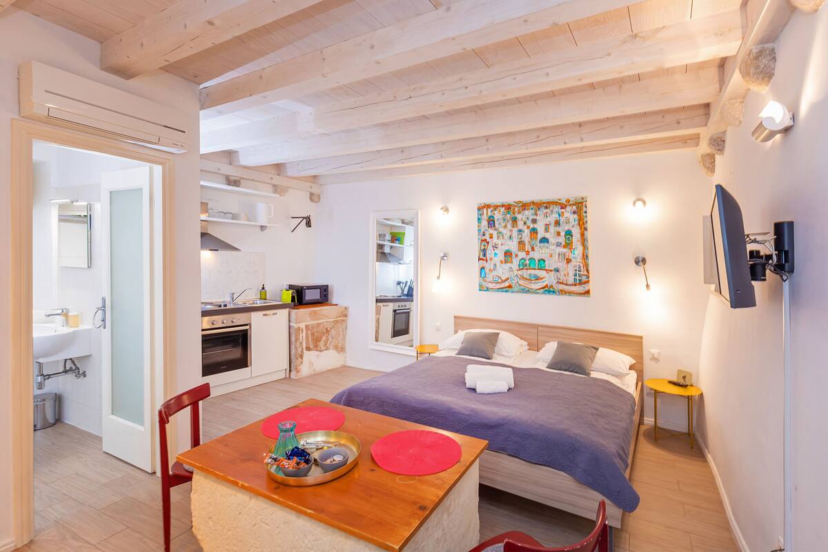 A compact hotel room with a bed, a small table with a tray, a kitchen area, and a bathroom.