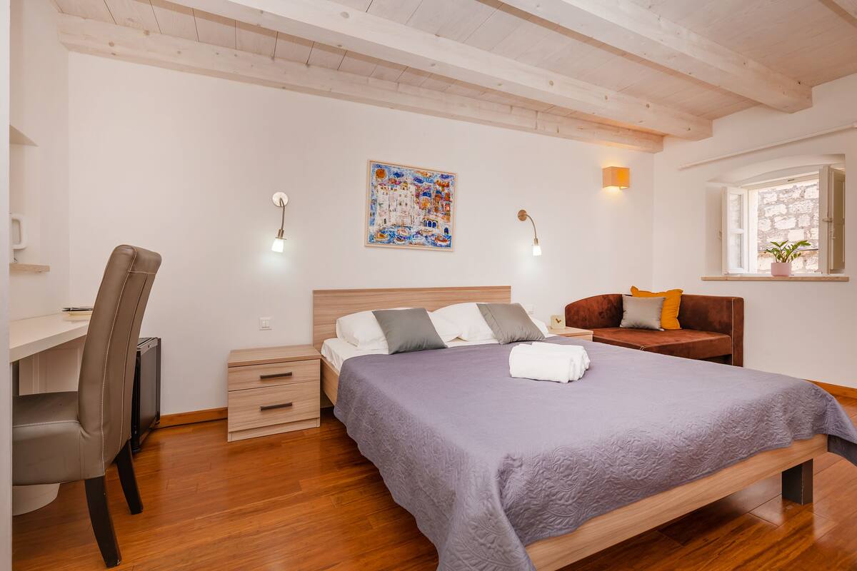 A bedroom with a bed, a desk, a chair, a nightstand, a window with a view, and a painting on the wall.