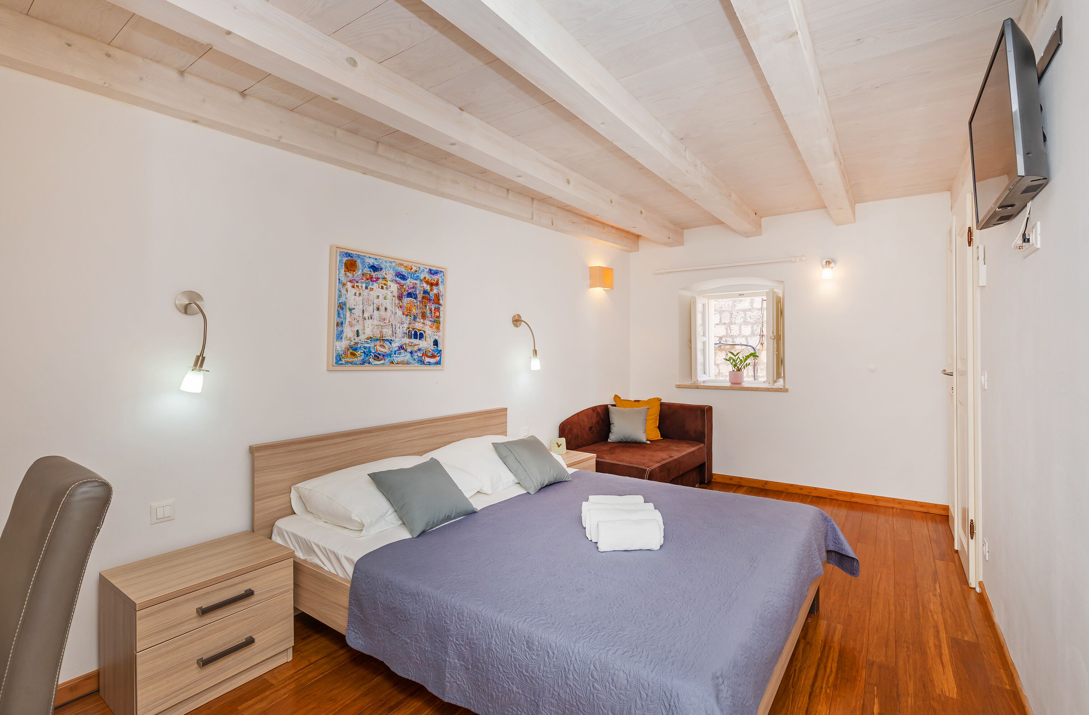 Un dormitorio con cama, escritorio, silla, ventana y un cuadro en la pared.