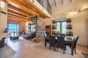 A spacious living area with a wooden ceiling, a dining table with chairs, a staircase, and a view through a large window.
