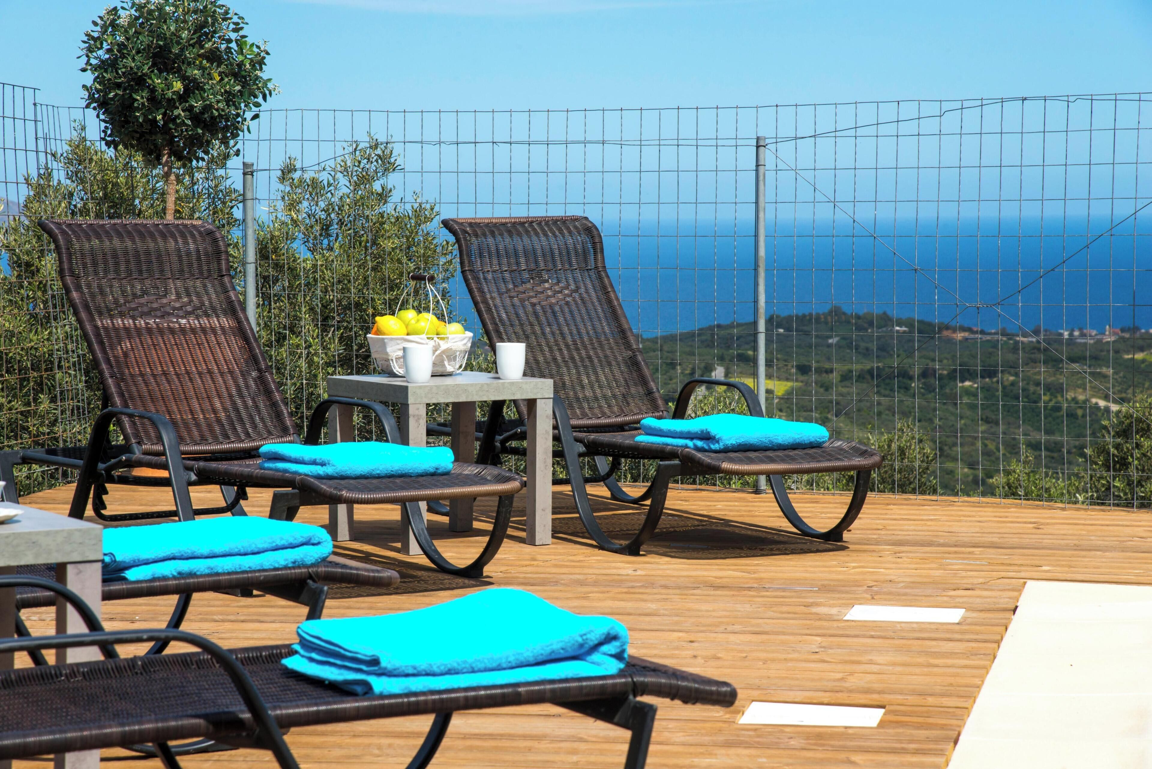 A deck with wicker lounge chairs, a table with a fruit bowl, and a view of the sea.