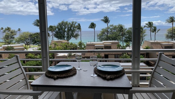 Varanda com mesa posta para dois, com vista para uma área costeira com edifícios e palmeiras.
