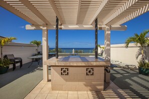 Un patio exterior con vista al océano, que incluye una pérgola, asientos y plantas en macetas.