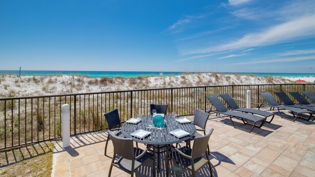 Eine Terrasse mit Tisch und Stühlen mit Blick auf einen Strand, Dünen und das Meer.