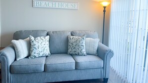 A grey sofa with floral cushions, a wall-mounted sign reading "BEACH HOUSE", and a floor lamp.