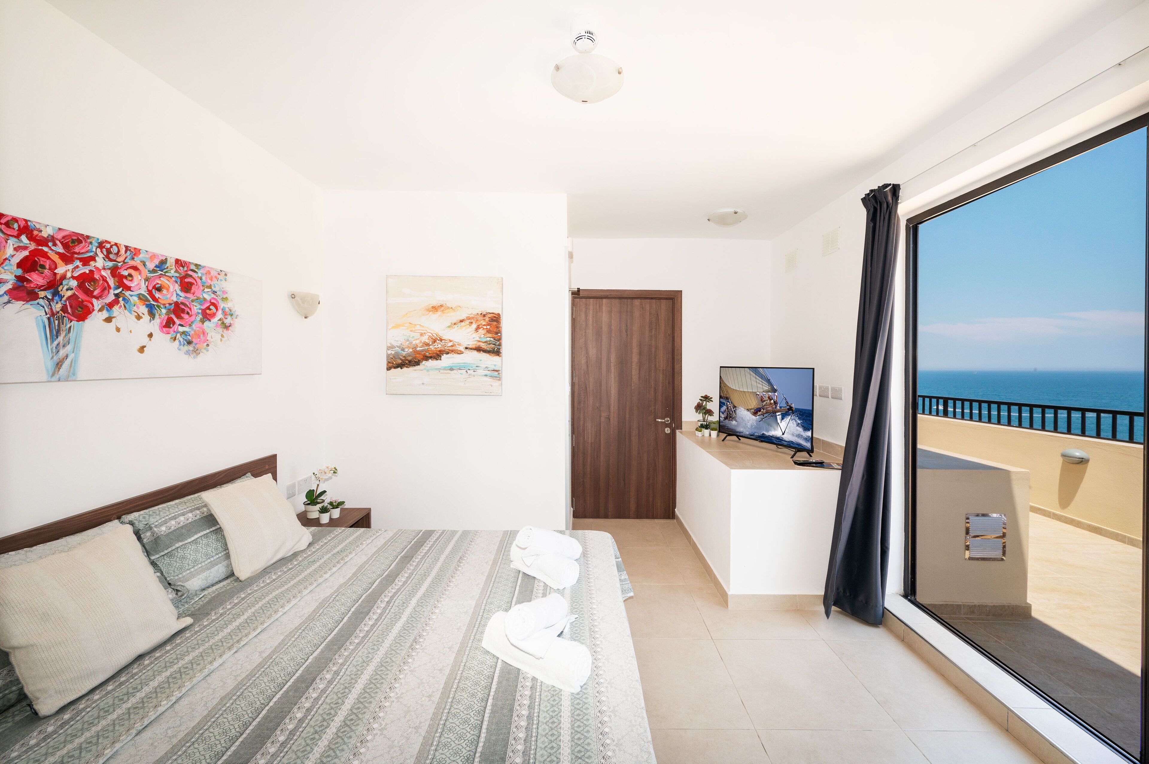 A bedroom with a bed, a desk, a TV, and a balcony with a view of the sea.