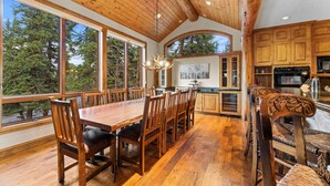 Un comedor espacioso con una larga mesa de madera y sillas, ventanas amplias que ofrecen vista al bosque, y una cocina con armarios de madera y electrodomésticos modernos.