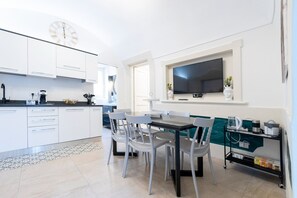 A modern kitchen with white cabinetry, a dining area with a table and chairs, a TV mounted on the wall, and a small shelf with kitchen appliances.
