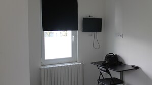 Une petite pièce avec une fenêtre, un radiateur, un bureau avec une chaise, un téléviseur fixé au mur et un sac noir posé sur le bureau.