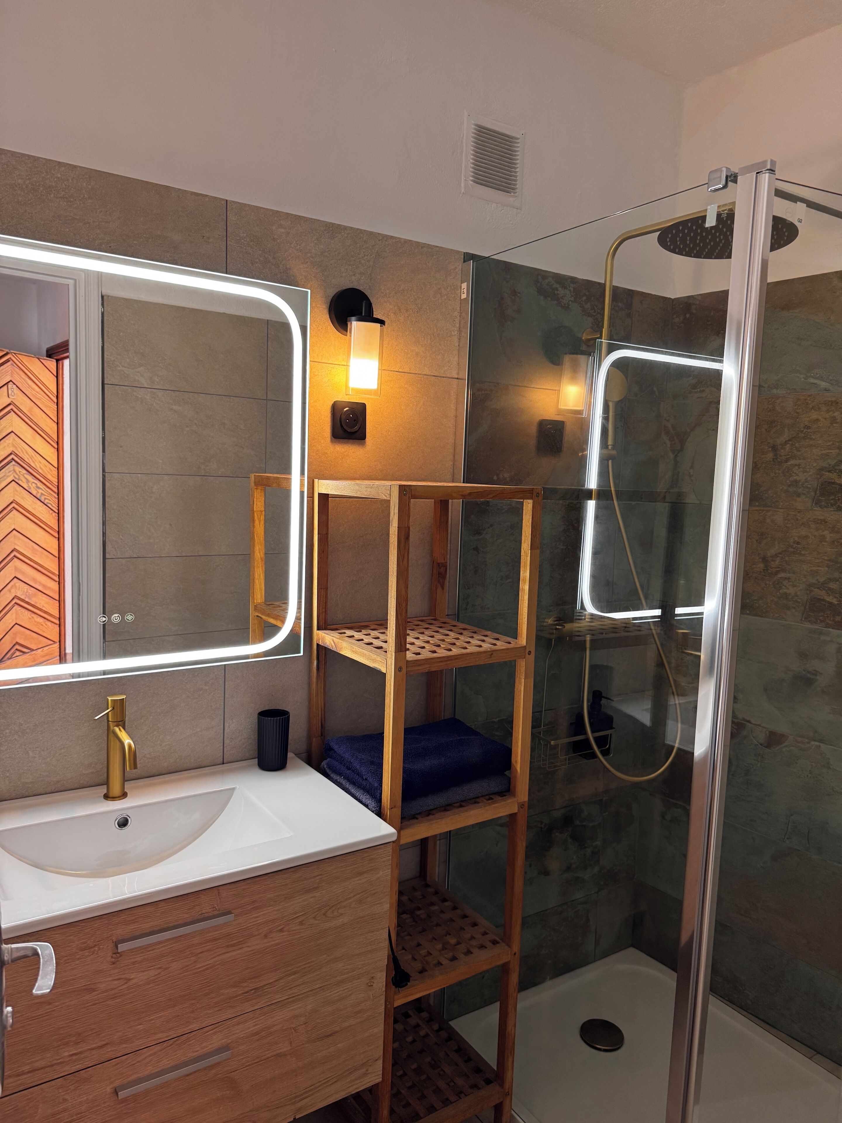 A bathroom with a white sink, a mirror with a light, a wooden shelf, and a glass shower enclosure.