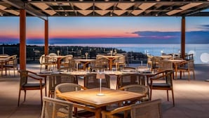 Un espacio de comedor al aire libre con mesas y sillas, vista al mar y un atardecer de fondo.