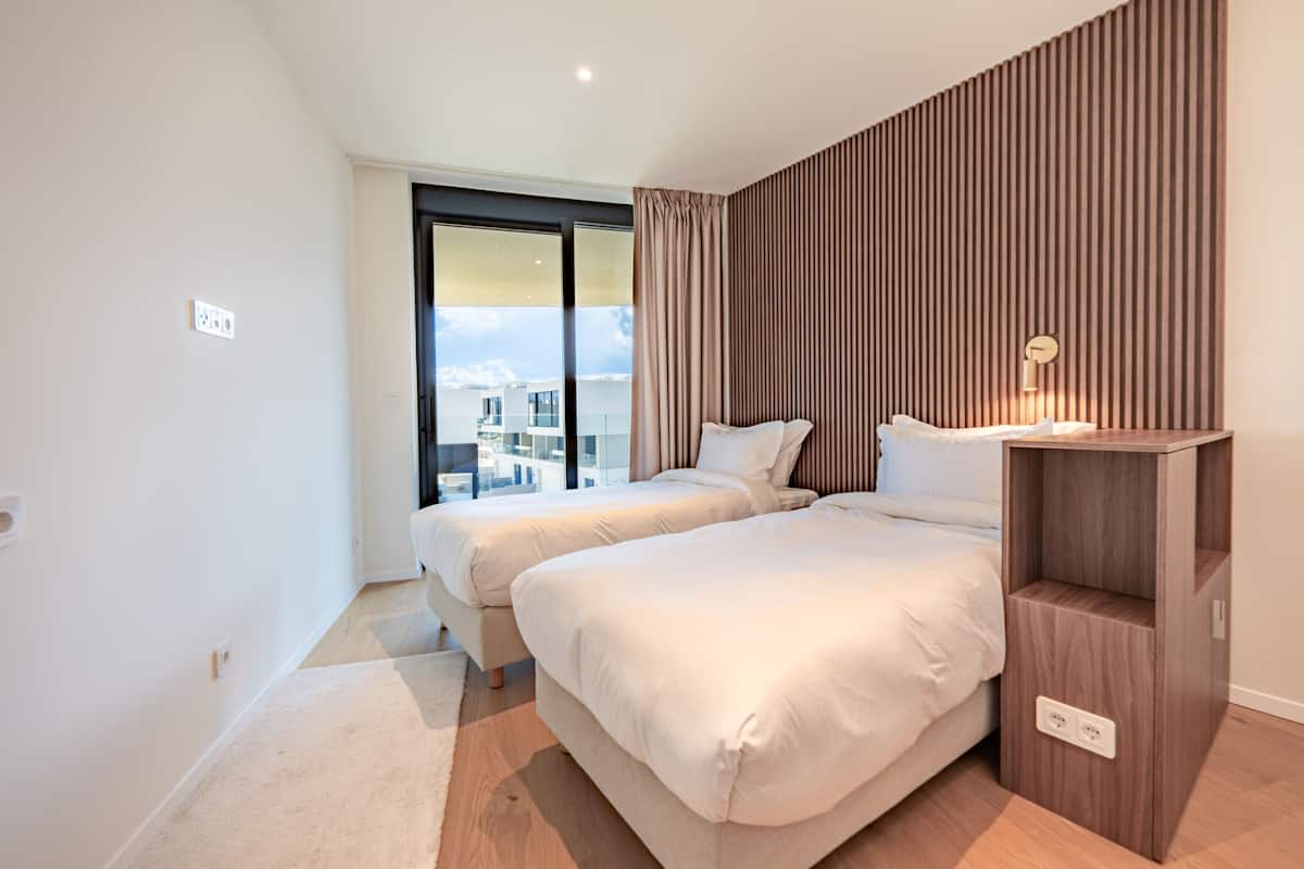 A modern hotel room with two beds, a wooden panel wall, a city view, and a bedside table with a lamp.