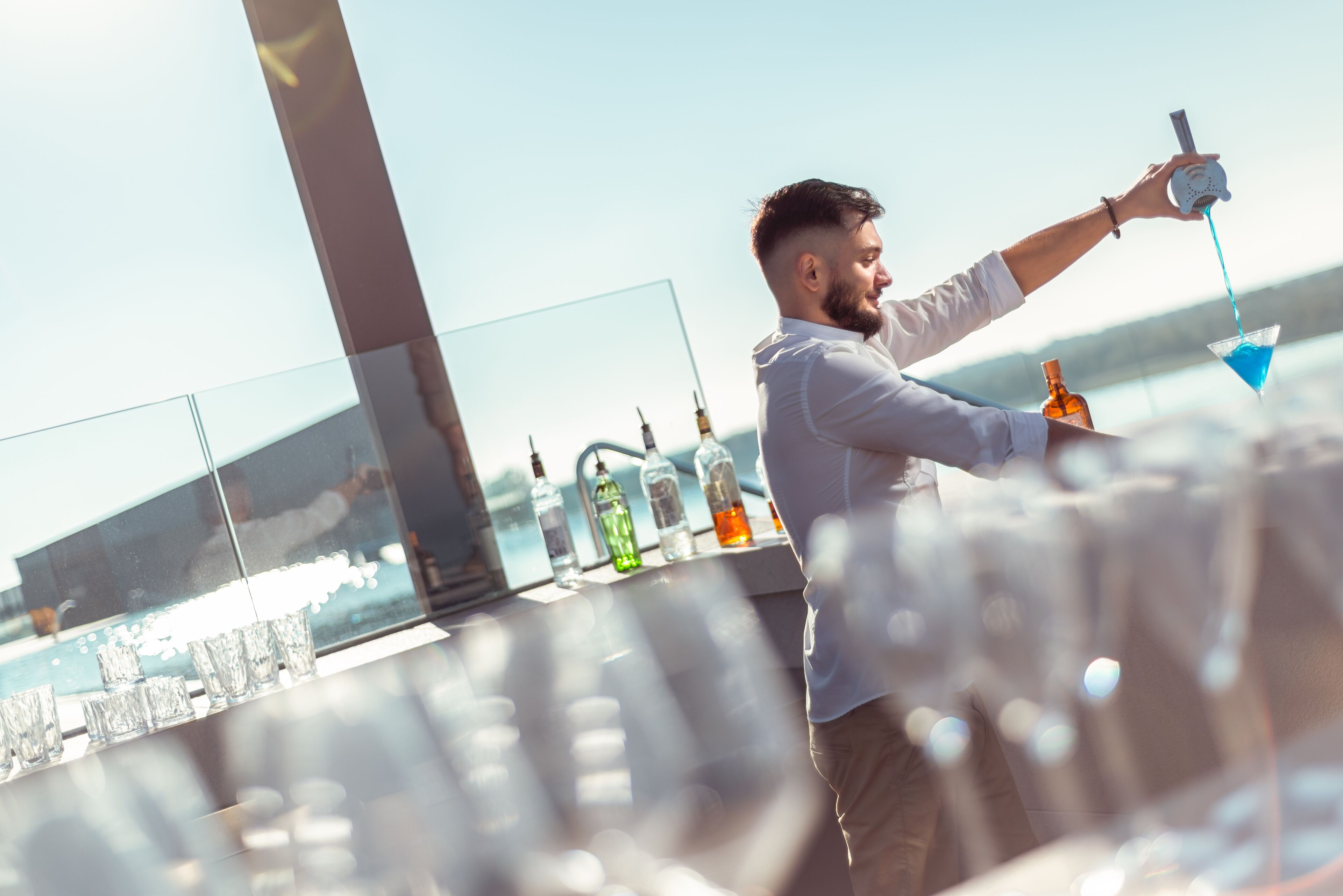 Una persona sirve una bebida en una copa de martini en un balcón con vista al mar y botellas de alcohol.