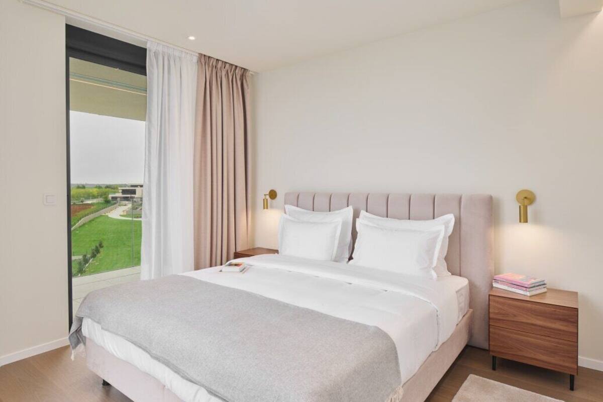 A bedroom with a large bed, a nightstand, a lamp, and a view of a green landscape through a window.