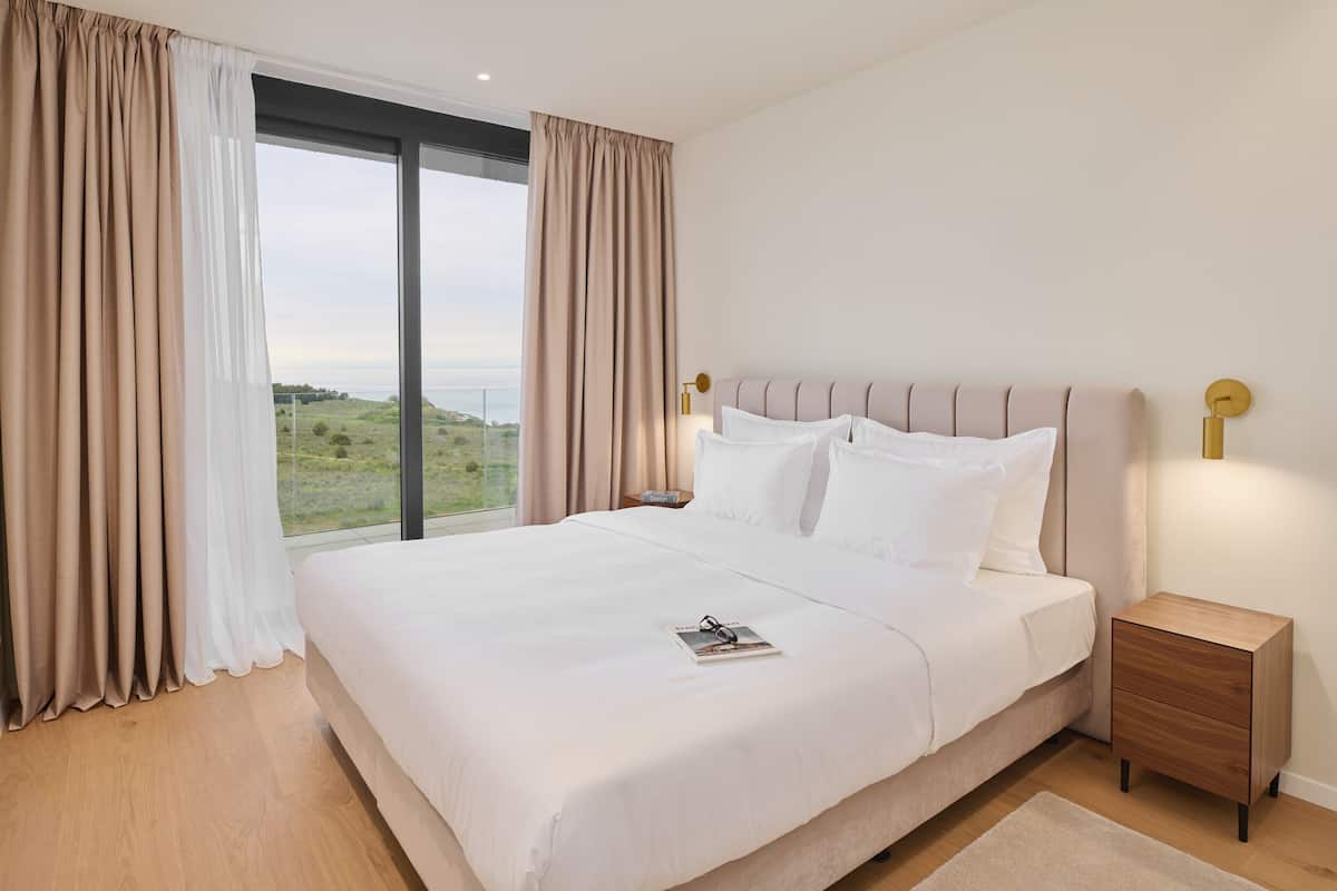 A neatly made bed with white linens, a bedside table with a lamp, and a view of the outdoors through a large window.