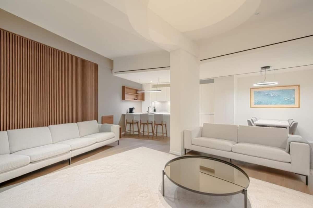 A modern living room with two white sofas, a glass coffee table, and a wooden accent wall. A kitchen area is visible in the background with bar stools.