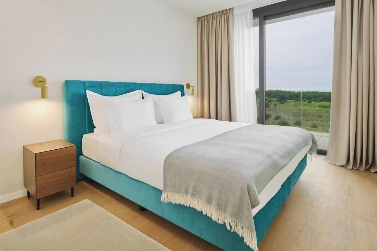 Una cama con cabecera color turquesa, ropa de cama blanca y vista a la vegetación por una ventana.