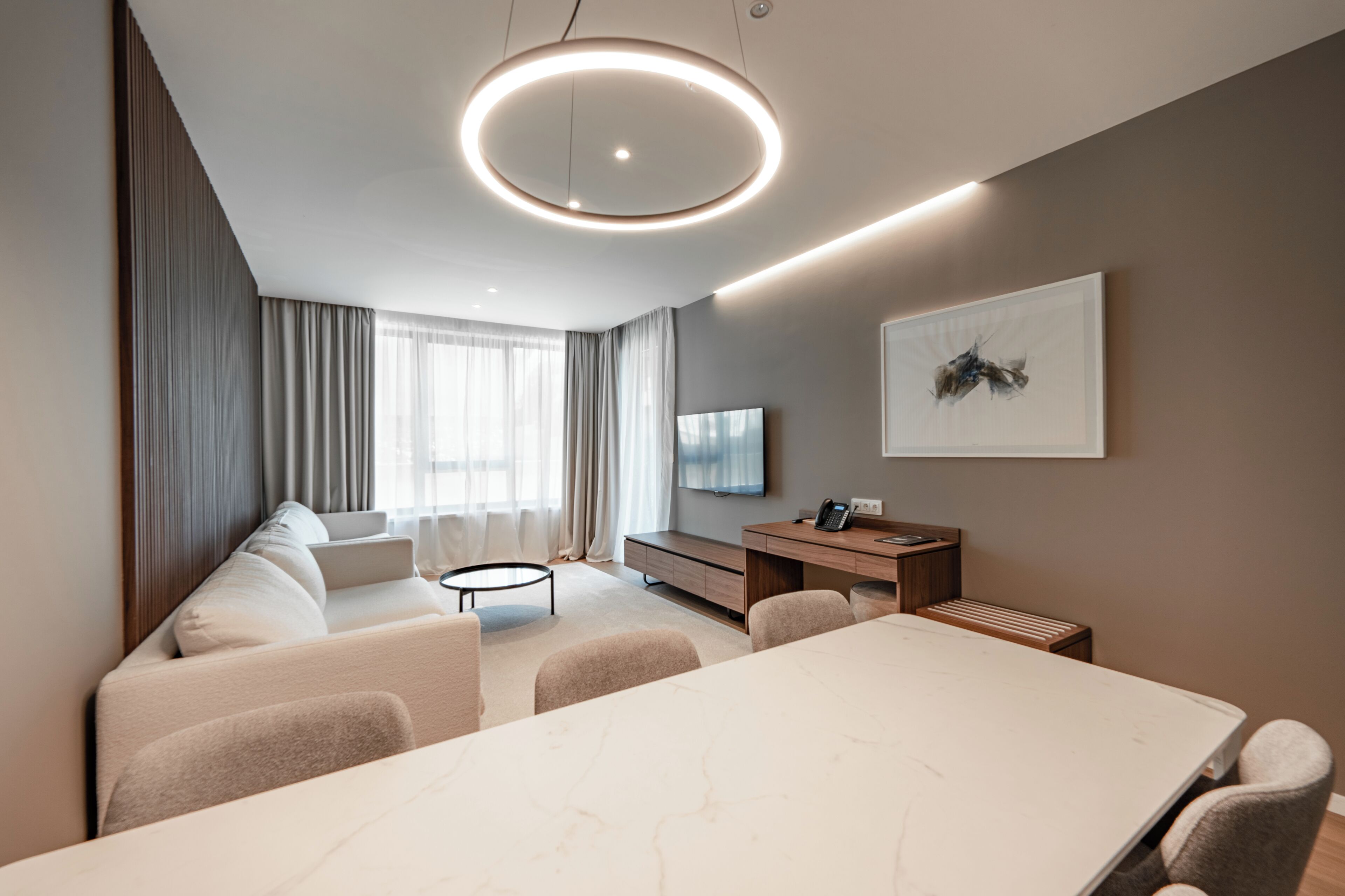A modern hotel room with a dining area, a sofa, a TV, and a painting on the wall.
