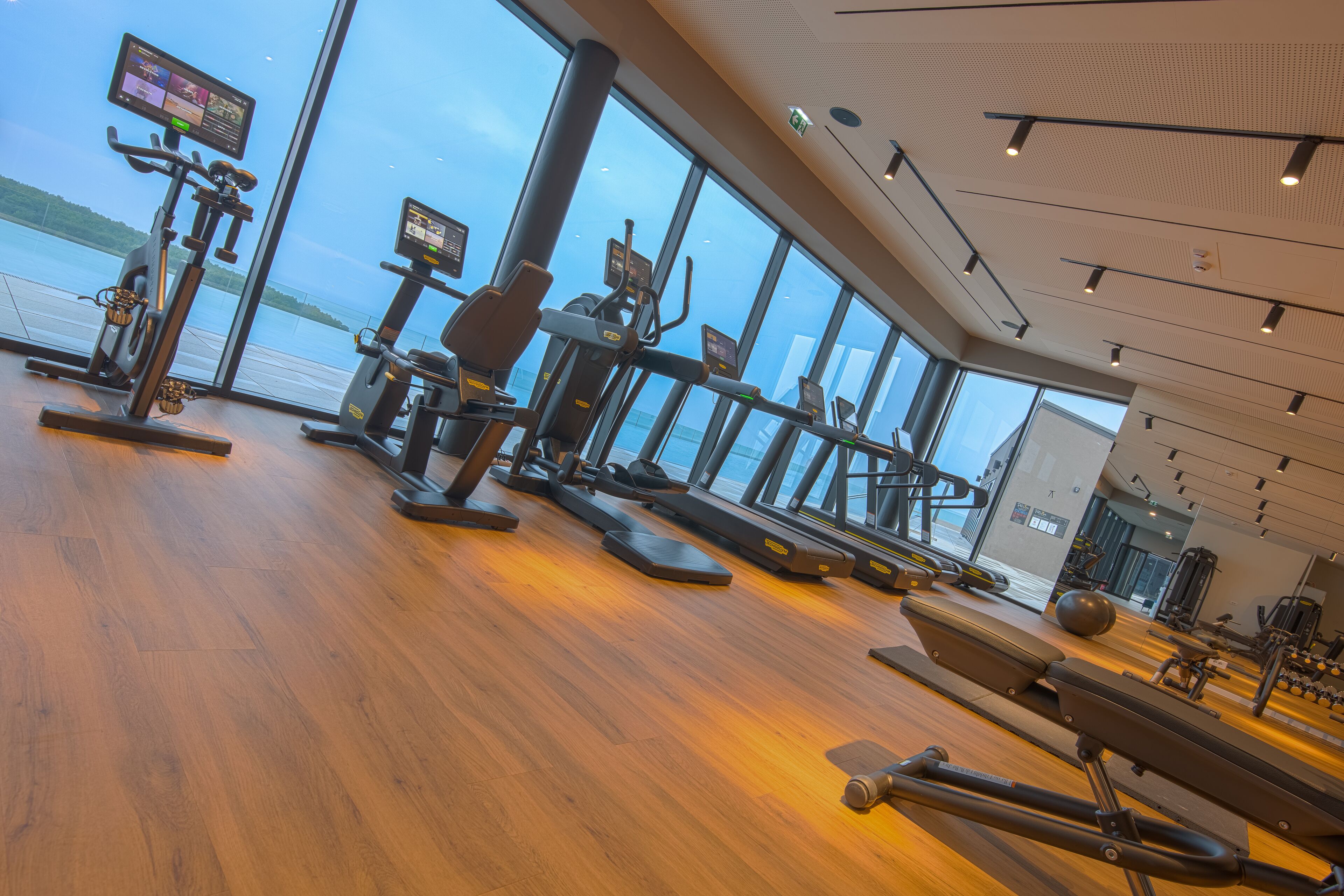 A gym with cardio machines, weight stations, and a large window overlooking a body of water.
